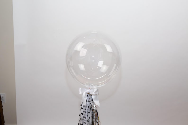 May include: A large, clear, round balloon with a white ribbon and a tassel of silver, black, and white confetti. The balloon is inflated and appears to be made of a transparent material. The balloon is a party decoration.