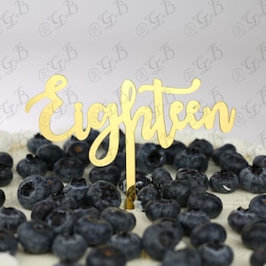 Könnte beinhalten: Ein weißer Kuchen mit Blaubeeren und einem goldenen Kuchentopper mit der Aufschrift "Eighteen".