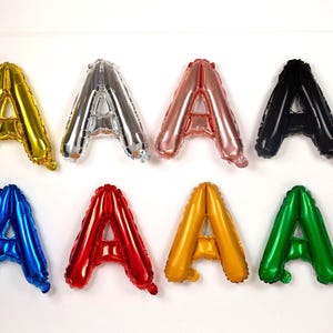 May include: Eight foil balloons shaped like the letter "A" in various colors: gold, silver, rose gold, black, blue, red, yellow, and green. The balloons are arranged against a white background.