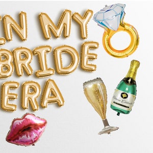 May include: Gold foil balloons spelling "In My Bride Era" with a pink lips balloon, a champagne flute balloon, a champagne bottle balloon, and a gold ring balloon with a white diamond.