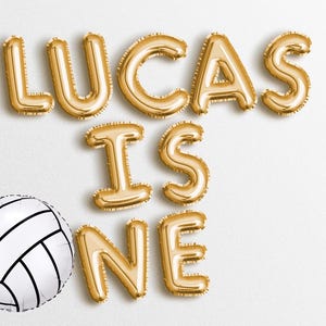 May include: Gold foil balloons spelling "Lucas is one" with a white and black volleyball.