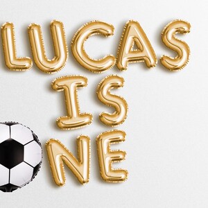 Op de afbeelding: Gouden folie letterballonnen die "Lucas is één" spellen met een zwart-witte voetbal.