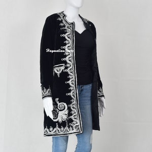 May include: A long black velvet jacket with white embroidered details. The jacket has a mandarin collar and a long sleeve. The embroidery is intricate and features a variety of floral and geometric patterns.