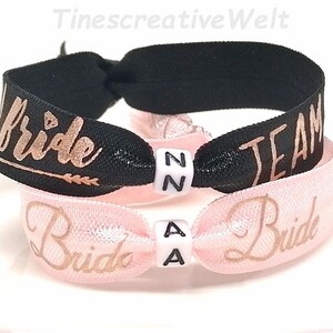Könnte beinhalten: Zwei elastische Haargummis, einer schwarz und einer rosa. Das schwarze Gummiband hat "Bride" und "TEAM" in goldener Schrift, und das rosa Gummiband hat "Bride". Jedes Gummiband hat einen kleinen weißen Würfel mit Buchstaben.