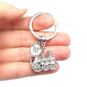 Könnte beinhalten: Silberfarbener Schlüsselanhänger mit einem Zugmotiv und einem runden Schlüsselring. Der Anhänger zeigt detaillierte Räder, einen Schornstein und ein rundes Emblem mit dem Buchstaben "W". Ein klassisches Accessoire.