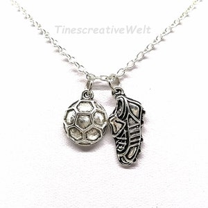 May include: A silver-colored necklace featuring two charms: a soccer ball and a soccer cleat. The soccer ball charm is a detailed, textured sphere. The cleat charm shows the side of a soccer shoe. The necklace chain is delicate.