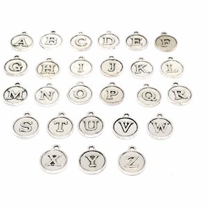 K&ouml;nnte beinhalten: Ein Satz von 26 silberfarbenen Metall-Anh&auml;ngern mit Buchstaben des Alphabets. Jeder Anh&auml;nger ist ein Kreis mit einem erhabenen Buchstaben in der Mitte.