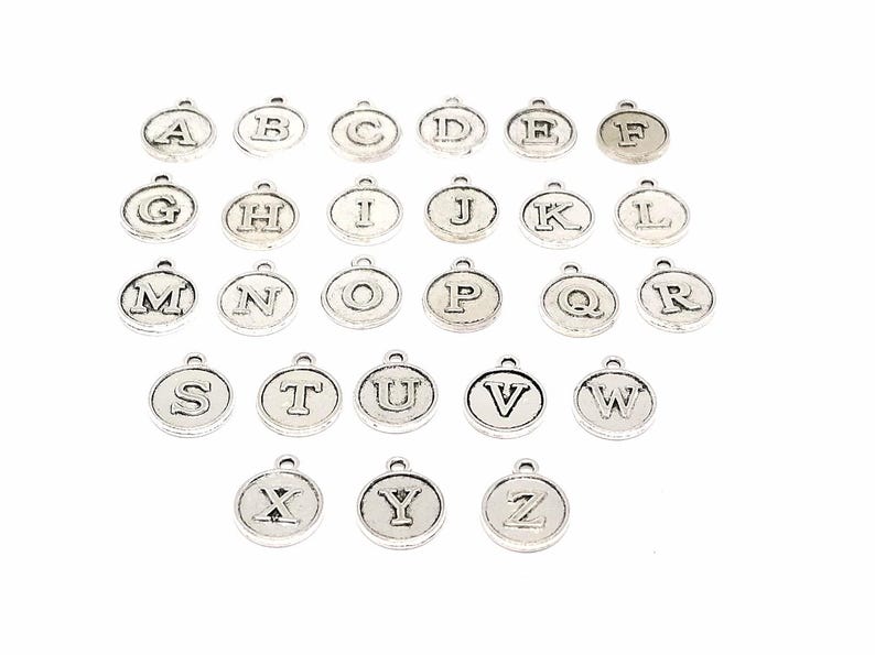 K&ouml;nnte beinhalten: Ein Satz von 26 silberfarbenen Metall-Anh&auml;ngern mit Buchstaben des Alphabets. Jeder Anh&auml;nger ist ein Kreis mit einem erhabenen Buchstaben in der Mitte.