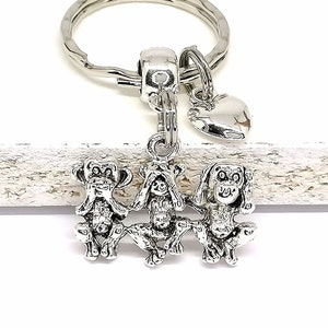Könnte beinhalten: Silberner Schlüsselanhänger mit drei Affen, einer bedeckt seine Augen, einer bedeckt seine Ohren und einer bedeckt seinen Mund. Die Affen sind miteinander verbunden und an einem silbernen Herzanhänger und einem silbernen Schlüsselring befestigt.