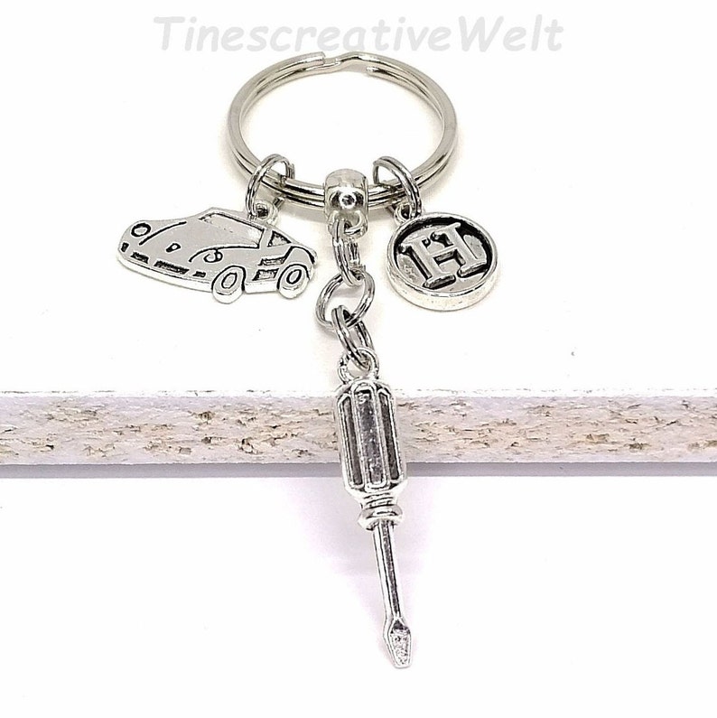 K&ouml;nnte beinhalten: Silberner Schl&uuml;sselanh&auml;nger mit einem Auto-Anh&auml;nger, einem Buchstaben-H-Anh&auml;nger und einem Schraubendreher-Anh&auml;nger. Der Schl&uuml;sselanh&auml;nger ist an einem silbernen Schl&uuml;sselring befestigt.