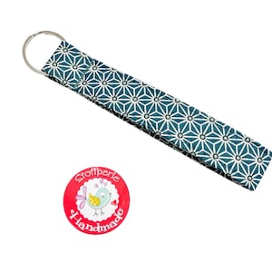 Könnte beinhalten: Ein türkisblauer und weiß gemusterter Stoffschlüsselanhänger mit einem silbernen Schlüsselring. Der Schlüsselanhänger hat einen runden Aufkleber mit einem rosa Vogel und dem Text "Stoffperle Handmade".