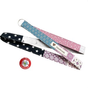 Könnte beinhalten: Zwei Schlüsselbänder aus Stoff mit Metallringen. Eines zeigt ein blaues geometrisches Muster und einen rosa gepunkteten Bereich mit einem Etikett mit der Aufschrift handmade. Das andere hat schwarzen Stoff mit weißen Sternen, Pusteblumen und violetten Mustern.