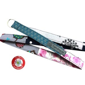 Könnte beinhalten: Ein Schlüsselband aus Stoff mit einem türkisfarbenen geometrischen Muster, einem grauen Abschnitt mit schwarzen Pusteblumen, einem marineblauen Abschnitt mit weißen Punkten und einem floralen Abschnitt mit rosa Rosen. Ein graues handmade-Etikett ist angebracht.