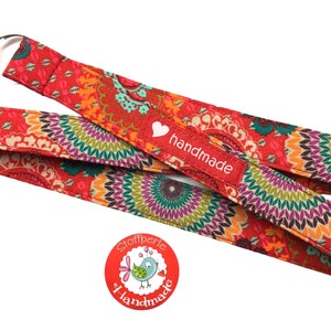 Könnte beinhalten: Roter und orangefarbener Stoffband mit einem bunten Mandala-Muster. Das Band hat einen silbernen Metallring und ein rotes Etikett mit der Aufschrift "handgefertigt".