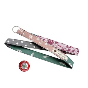 Könnte beinhalten: Drei Stoff-Lanyards mit verschiedenen Mustern: graue Sterne, weiße Blumen auf Rosa und weiße Federn auf Grün. Jedes Lanyard hat einen Metallring. Ein roter runder Aufkleber mit Text und einer Vogelillustration ist ebenfalls vorhanden.