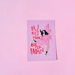 Könnte beinhalten: Eine rosa Postkarte mit dem deutschen Text "DU BIST DEIN ROTER FADEN" in Rot. Die Illustration zeigt eine Frau in einem rosa Kleid, mit schwarzen Haaren und schwarz-weißen Stiefeln. Der Hintergrund ist einfarbig rosa.