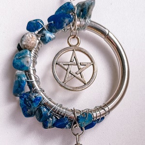 Könnte beinhalten: Ein silberner Metallring, um den blaue und weiße Steine gewickelt sind. Ein silberner Pentagramm-Anhänger hängt am Ring. Eine silberne Kette mit einem braunen Scheibenanhänger und einem silbernen Halbmondanhänger mit dem Text "Ich liebe dich bis zum Mond und zurück" hängt am Ring.