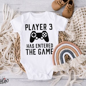 Könnte beinhalten: Weißer Baby Strampler mit schwarzem Text "Player 3 Has Entered The Game" und einer schwarzen Grafik einer Videospielkonsole.