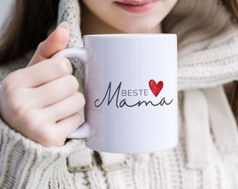 Könnte beinhalten: Weiße Keramiktasse mit einer schwarzen, kursiv geschriebenen Aufschrift "Beste Mama" mit einem roten Herzen über dem Wort "Mama".