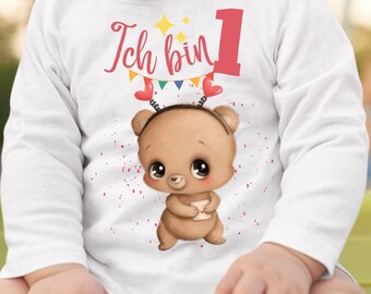 Könnte beinhalten: Ein weißer Langarm-Strampler mit dem Text "Ich bin 1" in rosa und gelb. Ein brauner Bär mit einem rosafarbenen Stirnband und einem rosafarbenen Herzen hält einen weißen Umschlag.