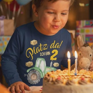 Könnte beinhalten: Ein Kind trägt ein marineblaues Langarmshirt mit einem grünen Traktor und dem Text "Platz da! ICH BIN 2". Das Kind pustet die Kerzen auf einer Geburtstagstorte aus. Ein Stofftier-Esel ist neben dem Kuchen.