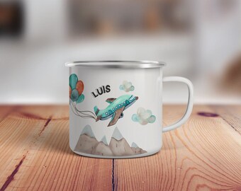 May include: White enamel mug with a blue airplane flying over brown mountains. The airplane has a blue and green design and is flying with a string of balloons. The mug has the name "LUIS" written on it.