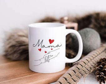 Könnte beinhalten: Weiße Keramiktasse mit einem schwarzen und roten Design. Die Tasse trägt den Schriftzug "Mama" mit einem roten Herzen über dem Wort. Es gibt eine schwarze Linienzeichnung von zwei Händen, die sich halten, mit drei roten Herzen über den Händen.