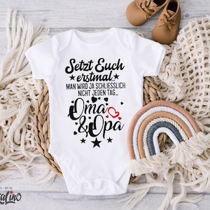 Könnte beinhalten: Weißer Baby Strampler mit schwarzem Text, der "Setzt Euch erstmal... Man wird ja schliesslich nicht jeden Tag... Oma & Opa" mit einem roten Herzen und Sternen lautet.