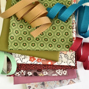 Könnte beinhalten: Eine Sammlung von Stoffstücken und Bändern in verschiedenen Farben. Die Stoffe umfassen grüne Blumenmuster, Blumenmuster und unifarbene Stoffe. Bänder sind beige, türkis, rot und mintgrün. Ideal für Näh- und Bastelprojekte.