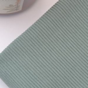Puede incluir: Una tela de color verde salvia que presenta una textura acanalada vertical distintiva. El material est&aacute; doblado, mostrando las l&iacute;neas paralelas consistentes del patr&oacute;n de tejido en toda la superficie. El color es un tono verde terroso.