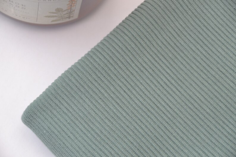 Puede incluir: Una tela de color verde salvia que presenta una textura acanalada vertical distintiva. El material est&aacute; doblado, mostrando las l&iacute;neas paralelas consistentes del patr&oacute;n de tejido en toda la superficie. El color es un tono verde terroso.