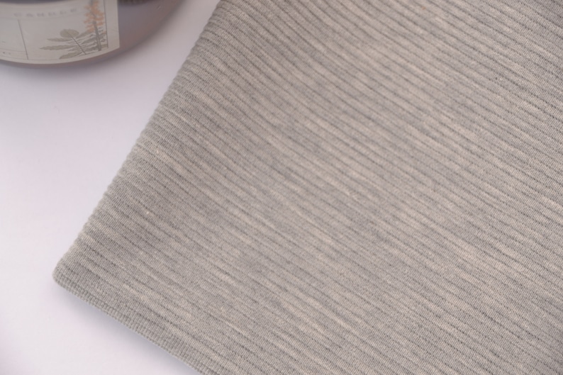 Puede incluir: Una pieza de tela gris claro con una textura finamente acanalada. El material muestra un patr&oacute;n de tejido consistente con l&iacute;neas verticales que recorren la superficie, creando una apariencia suave y uniforme en todo el tejido visible.
