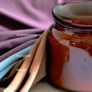 K&ouml;nnte beinhalten: Ein dunkel bernsteinfarbenes Glasgef&auml;&szlig; mit Deckel neben drei gefalteten Stoffmustern. Die Stofffarben sind als dunkelmauve, dustyblue und nougat beschriftet. Das Glasgef&auml;&szlig; hat eine glatte, reflektierende Oberfl&auml;che und eine zylindrische Form.