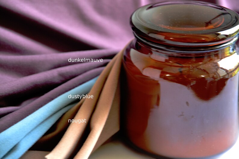 K&ouml;nnte beinhalten: Ein dunkel bernsteinfarbenes Glasgef&auml;&szlig; mit Deckel neben drei gefalteten Stoffmustern. Die Stofffarben sind als dunkelmauve, dustyblue und nougat beschriftet. Das Glasgef&auml;&szlig; hat eine glatte, reflektierende Oberfl&auml;che und eine zylindrische Form.