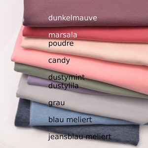 K&ouml;nnte beinhalten: Ein Stapel gefalteter Stoffe in verschiedenen Farben, darunter dunkelmauve, marsala, poudre, candy, dustymint, dustylila, grau, blau meliert und jeansblau meliert, wobei die Farbnamen auf jeder entsprechenden Stoffschicht aufgedruckt sind.