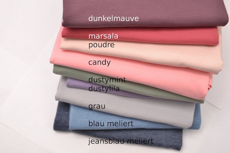 K&ouml;nnte beinhalten: Ein Stapel gefalteter Stoffe in verschiedenen Farben, darunter dunkelmauve, marsala, poudre, candy, dustymint, dustylila, grau, blau meliert und jeansblau meliert, wobei die Farbnamen auf jeder entsprechenden Stoffschicht aufgedruckt sind.
