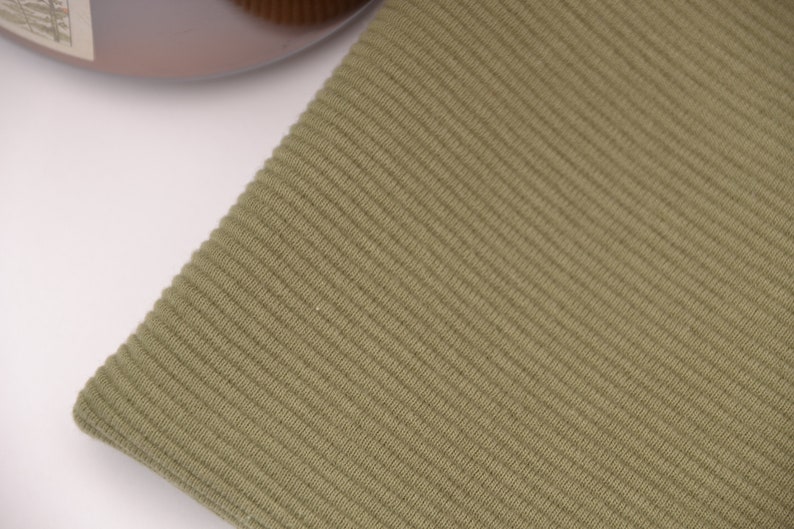 Puede incluir: Un trozo de tela de color verde oliva con una textura acanalada distintiva. El material muestra un patr&oacute;n vertical consistente de l&iacute;neas en relieve en toda su superficie, creando una apariencia uniforme y t&aacute;ctil en toda la secci&oacute;n visible del tejido.