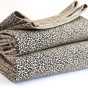 Puede incluir: Dos piezas de tela dobladas apiladas una encima de la otra. El tejido presenta un patrón repetitivo de estampado de leopardo negro sobre un fondo beige claro. Los bordes de la tela son visibles, mostrando una estructura plegada.
