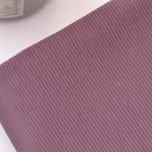 Puede incluir: Una tela de color malva que presenta una textura acanalada vertical distintiva. El material muestra un patr&oacute;n de tejido fino y consistente en toda su superficie, caracterizado por l&iacute;neas paralelas en relieve que crean una profundidad sutil.