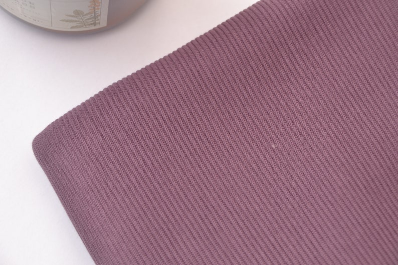 Puede incluir: Una tela de color malva que presenta una textura acanalada vertical distintiva. El material muestra un patr&oacute;n de tejido fino y consistente en toda su superficie, caracterizado por l&iacute;neas paralelas en relieve que crean una profundidad sutil.