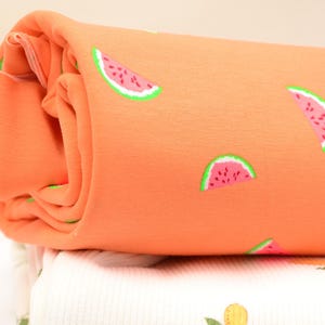 Könnte beinhalten: Gerollter orangefarbener Stoff mit einem sich wiederholenden Muster aus Wassermelonenscheiben. Die Wassermelonen haben rosa Fruchtfleisch, grüne Schalen und schwarze Kerne. Der Stoff ist wahrscheinlich Baumwolle oder ein ähnliches Material, geeignet für Kleidung oder Heimdekoration.