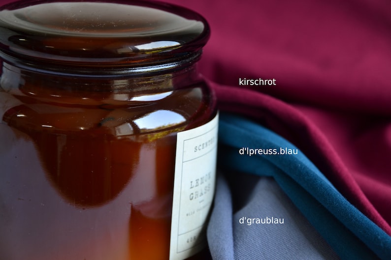 K&ouml;nnte beinhalten: Ein dunkel bernsteinfarbenes Glasgef&auml;&szlig; mit Deckel und einem wei&szlig;en Etikett mit der Aufschrift LEMON GRASS, neben drei gefalteten Stoffmustern in Kirschrot, Preu&szlig;ischblau und Graublau, mit dem Text kirschrot, d'lpreuss.blau und d'graublau.