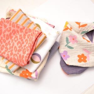 Puede incluir: Una pila de telas de muselina dobladas con varios patrones, incluyendo un estampado de leopardo coral, rayas naranjas y grises, y motivos florales coloridos, junto a un babero triangular con fondo crema y estampado floral.