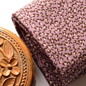 Puede incluir: Una pieza de tela doblada con fondo morado y un estampado de leopardo en marrón y crema, situada junto a un recipiente redondo de madera con un diseño floral tallado en la tapa y un pomo central como tirador.