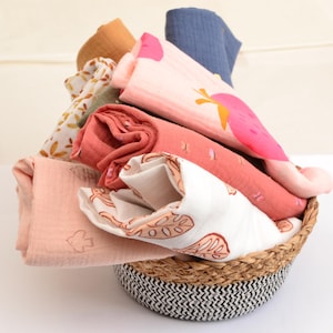 Könnte beinhalten: Ein geflochtener Korb gefüllt mit einem Stapel bunter Baby-Swaddles. Die Swaddles sind aus weichem, atmungsaktivem Stoff gefertigt und haben verschiedene Muster, darunter florale, geometrische und abstrakte Designs.