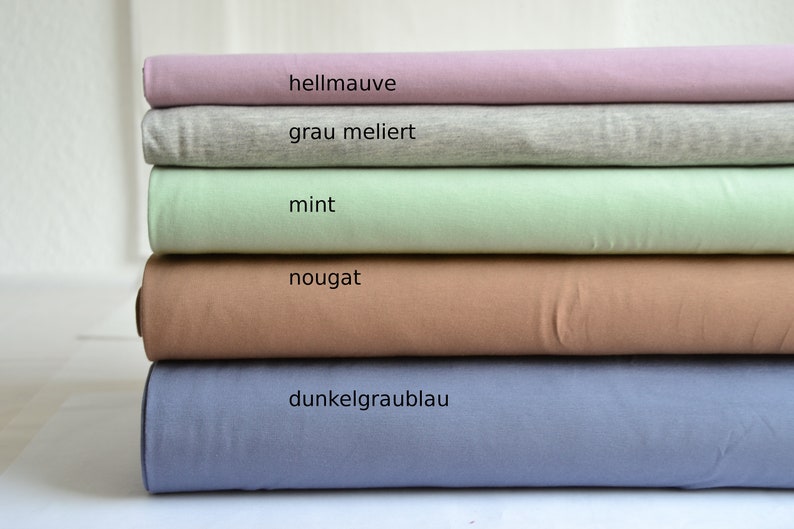 K&ouml;nnte beinhalten: Ein Stapel aus f&uuml;nf gefalteten Stoffballen in Unifarben. Die Farben von oben nach unten sind als hellmauve, grau meliert, mint, nougat und dunkelgraublau beschriftet. Jeder Stoffballen ist ordentlich horizontal &uuml;bereinander geschichtet.