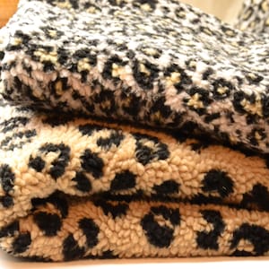 Puede incluir: Dos piezas de tela texturizada dobladas con un patrón de leopardo. La pieza superior muestra un diseño de manchas negras, blancas y grises, mientras que la pieza inferior presenta un estampado de leopardo negro y beige sobre una base crema.