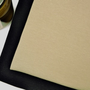 Puede incluir: Dos piezas rectangulares de tela acanalada superpuestas. La pieza superior es de color beige claro, mientras que la pieza inferior es negra. Ambas telas presentan una textura acanalada vertical fina y uniforme en toda la superficie.