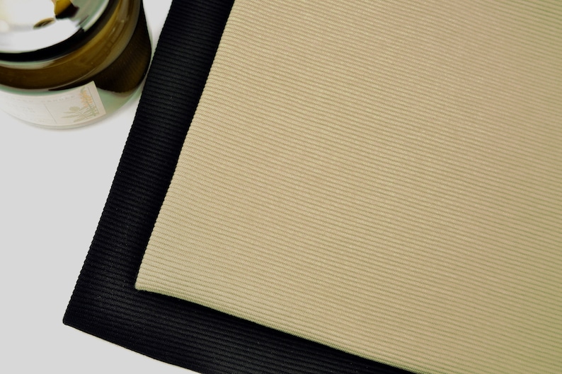 Puede incluir: Dos piezas rectangulares de tela acanalada superpuestas. La pieza superior es de color beige claro, mientras que la pieza inferior es negra. Ambas telas presentan una textura acanalada vertical fina y uniforme en toda la superficie.