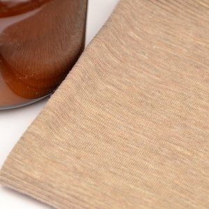 Puede incluir: Una pieza de tela de color tostado claro doblada con una textura fina y jaspeada. Junto a la tela hay una parte de un recipiente de vidrio lleno de un l&iacute;quido &aacute;mbar oscuro, con una etiqueta rectangular que muestra una ilustraci&oacute;n bot&aacute;nica de flores amarillas.